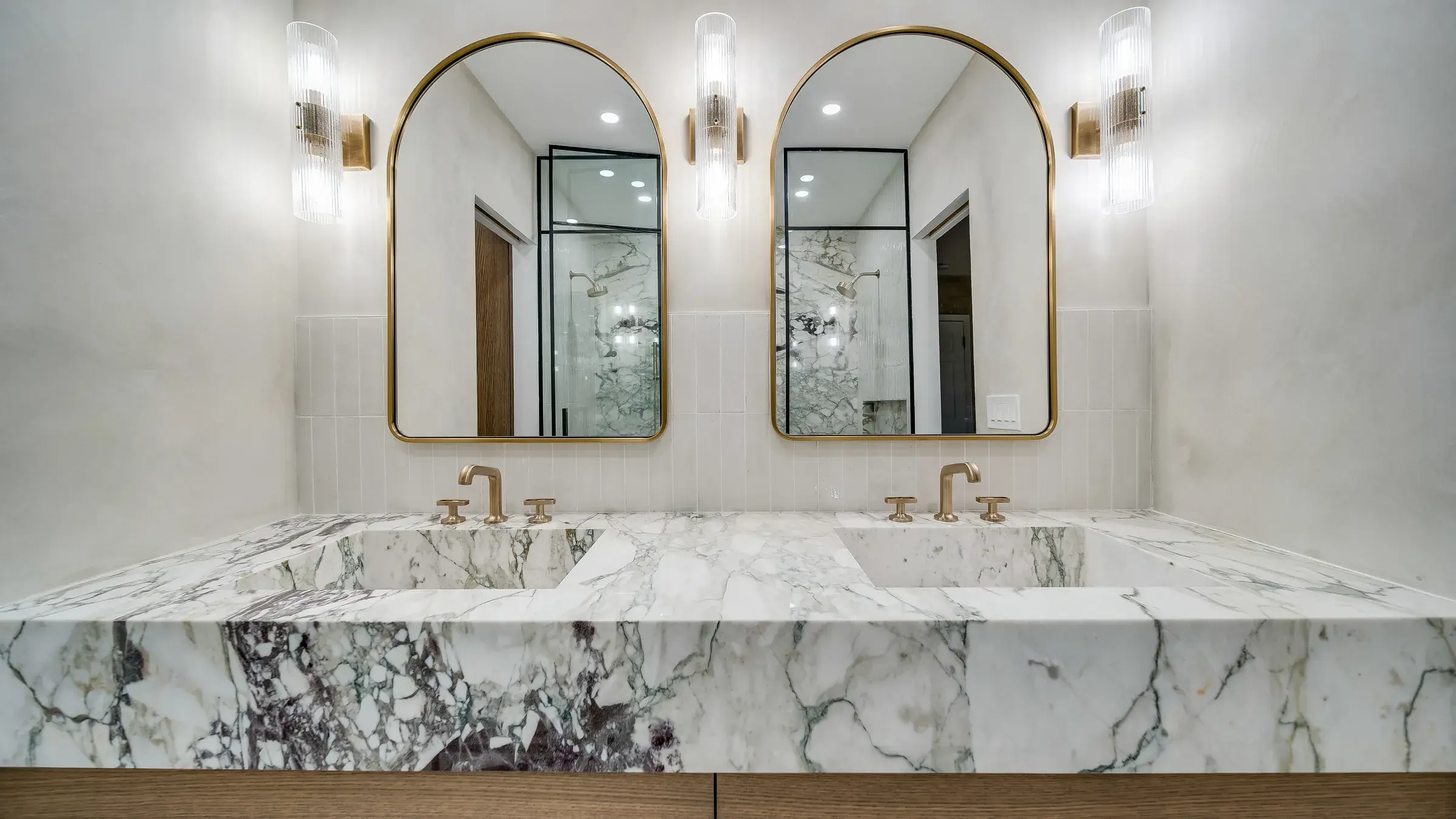 Double vanity: marble slab with integrated sinks, brass faucets, twin arched mirrors and sconces