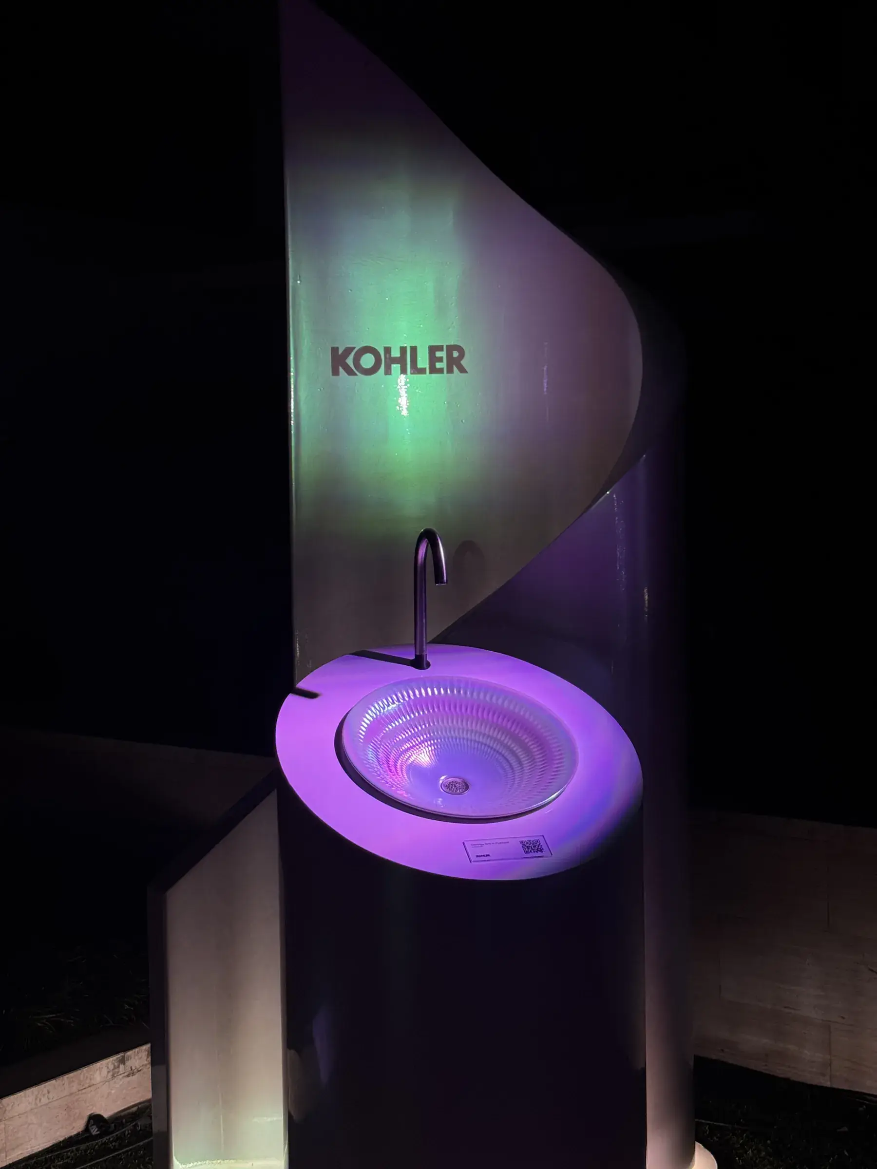 Cylindrical product pedestal with curved metal backing panel, KOHLER logo and lit pearlized sink