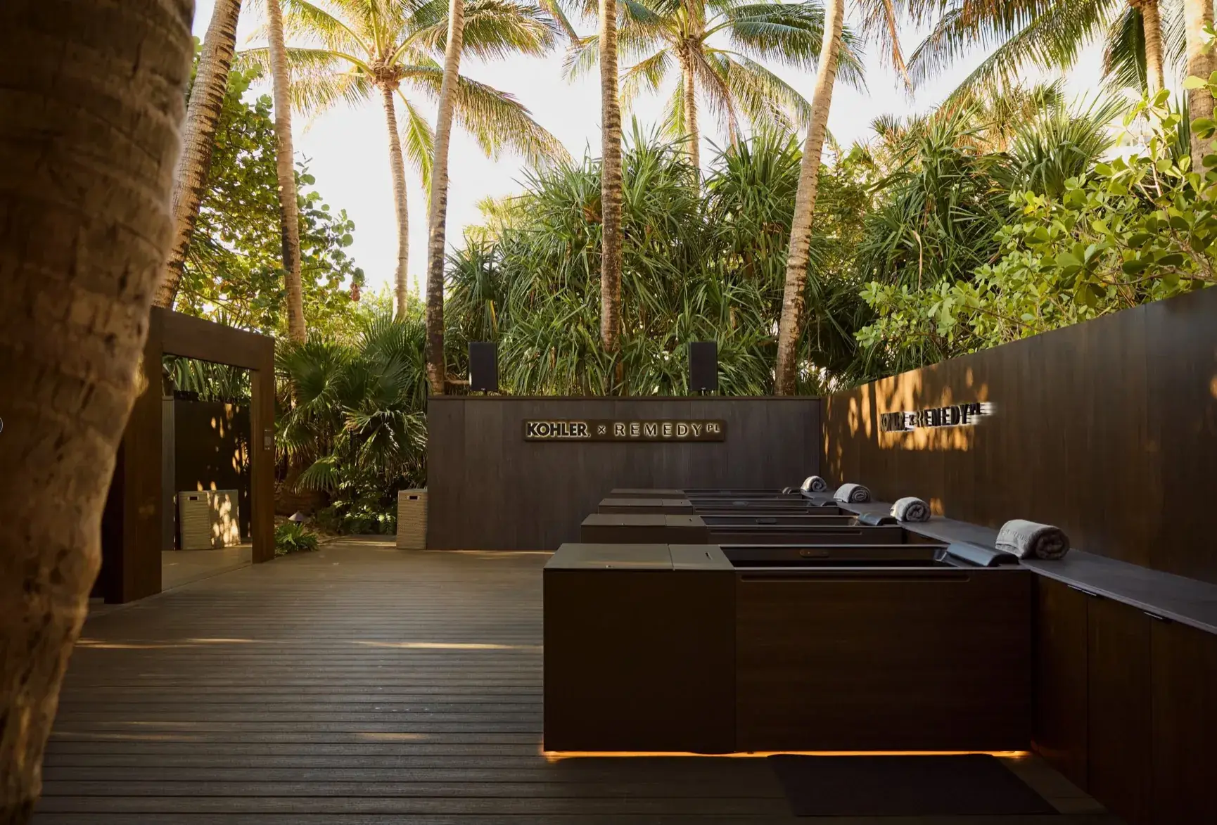 KOHLER × REMEDY cold-plunge deck at golden hour: wood decking, timber-clad tubs with LED underglow, shower pergola, and signage