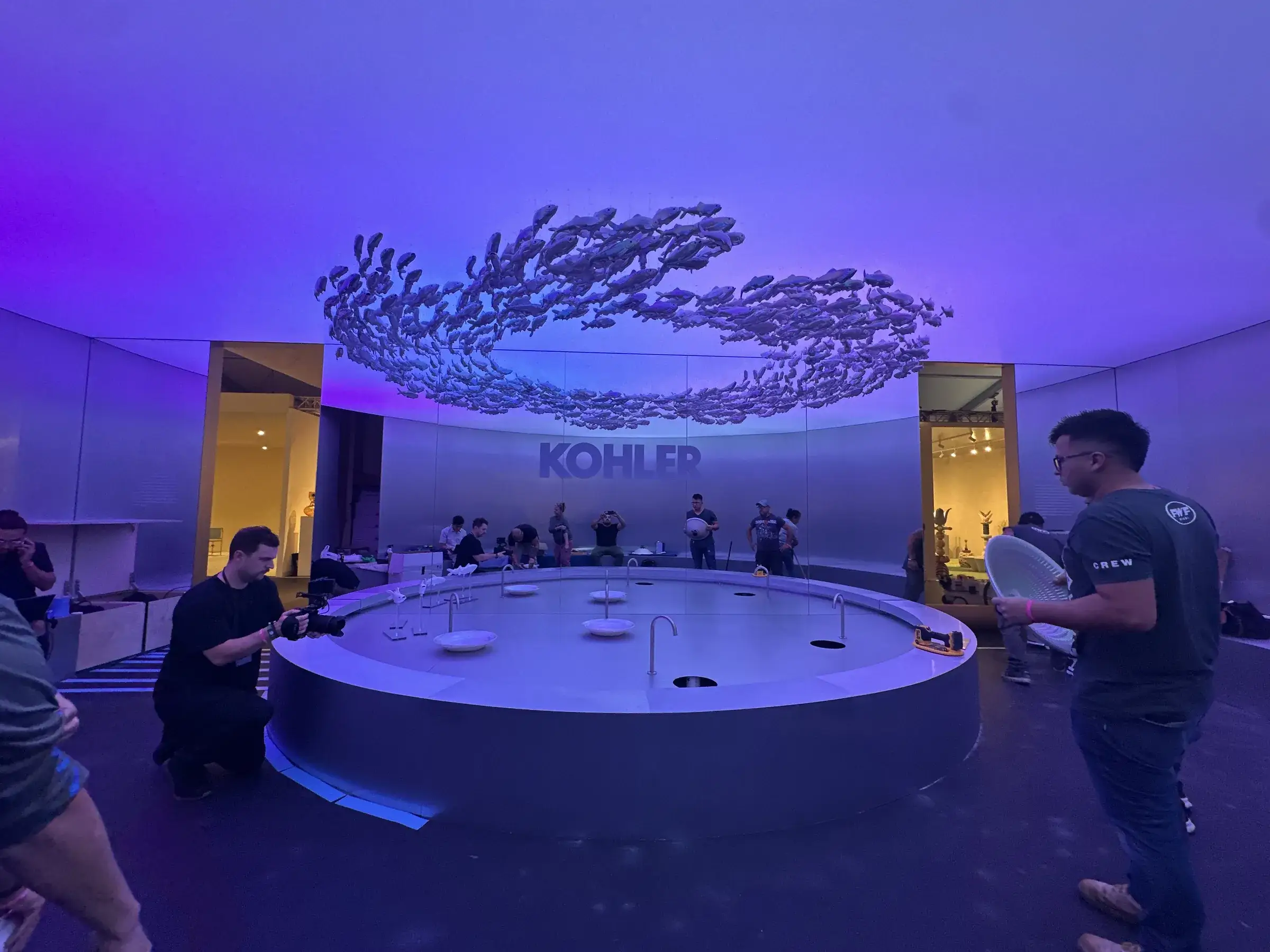 Kohler booth at Design Miami 2025: KOHLER logo wall, suspended fish installation, and circular basin under purple and blue lighting