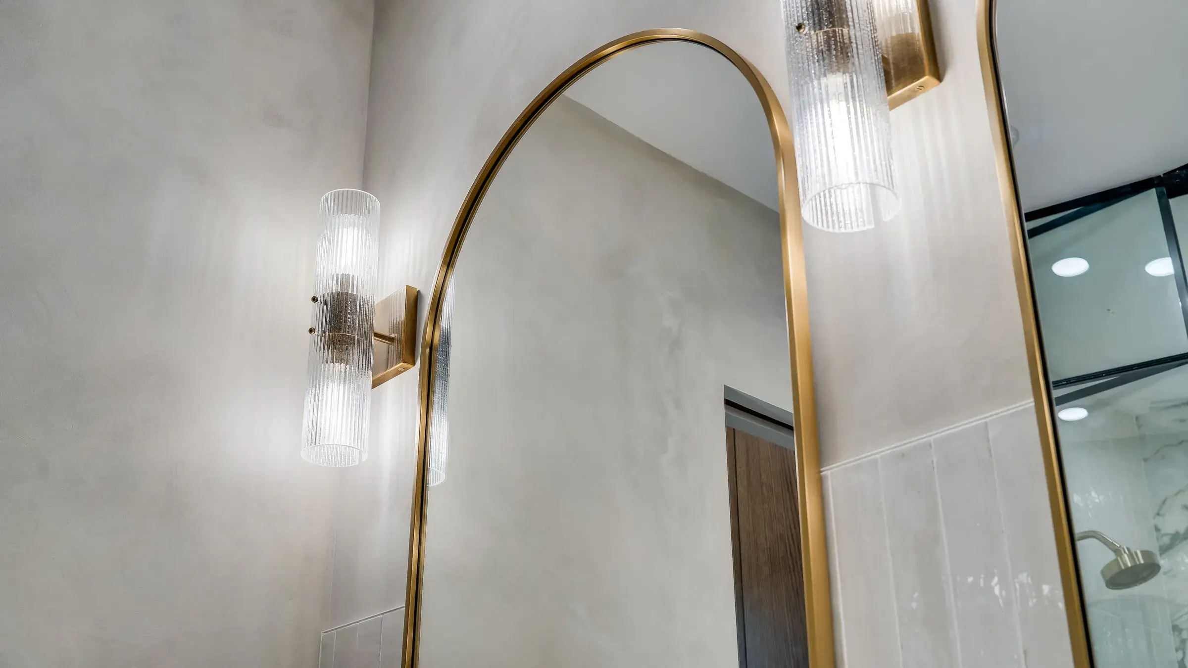 Arched brass mirror edge with ribbed-glass sconce against the plaster wall