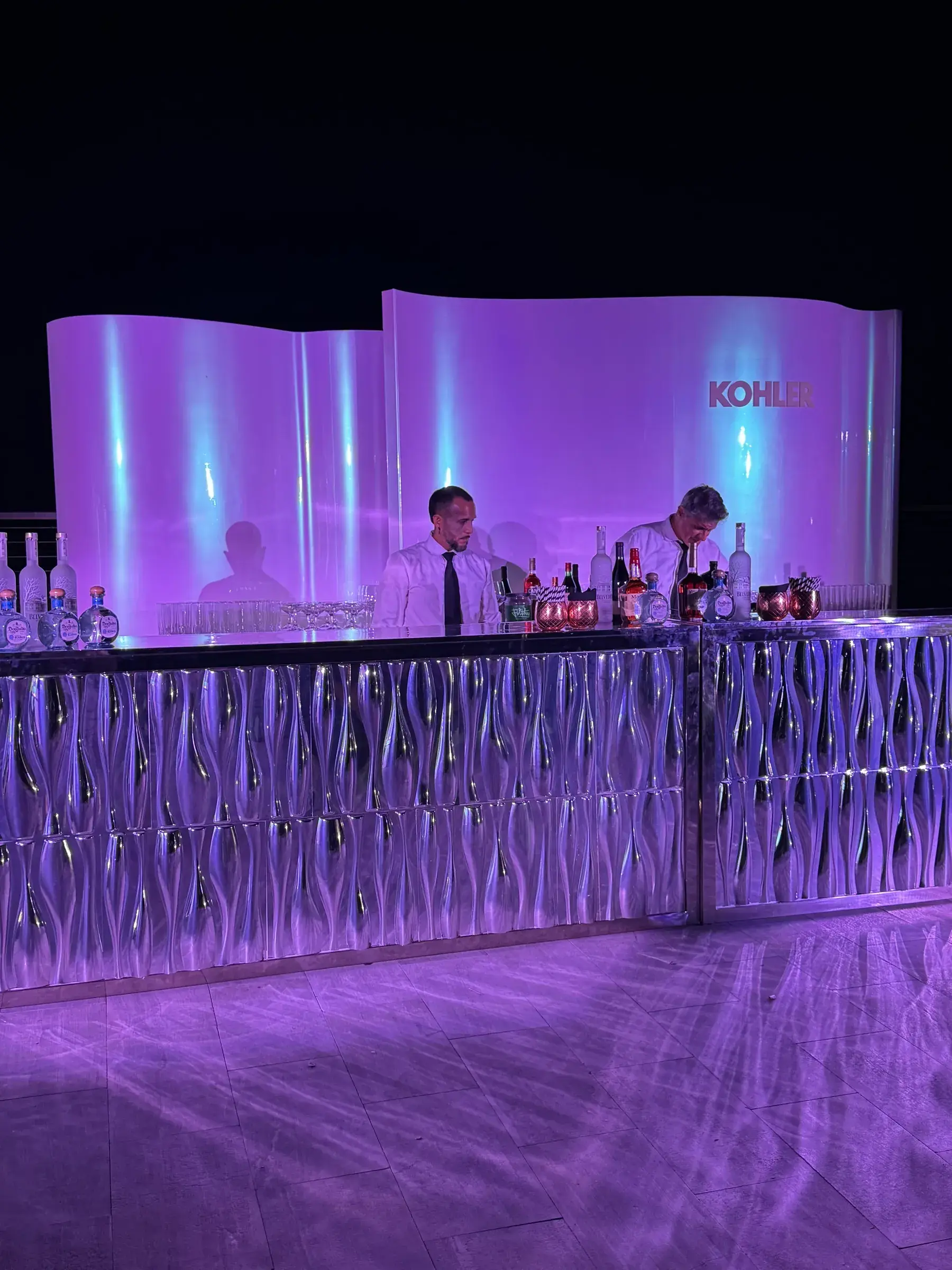 Curved KOHLER backdrop panels and ripple-textured mirror bar front under purple event lighting